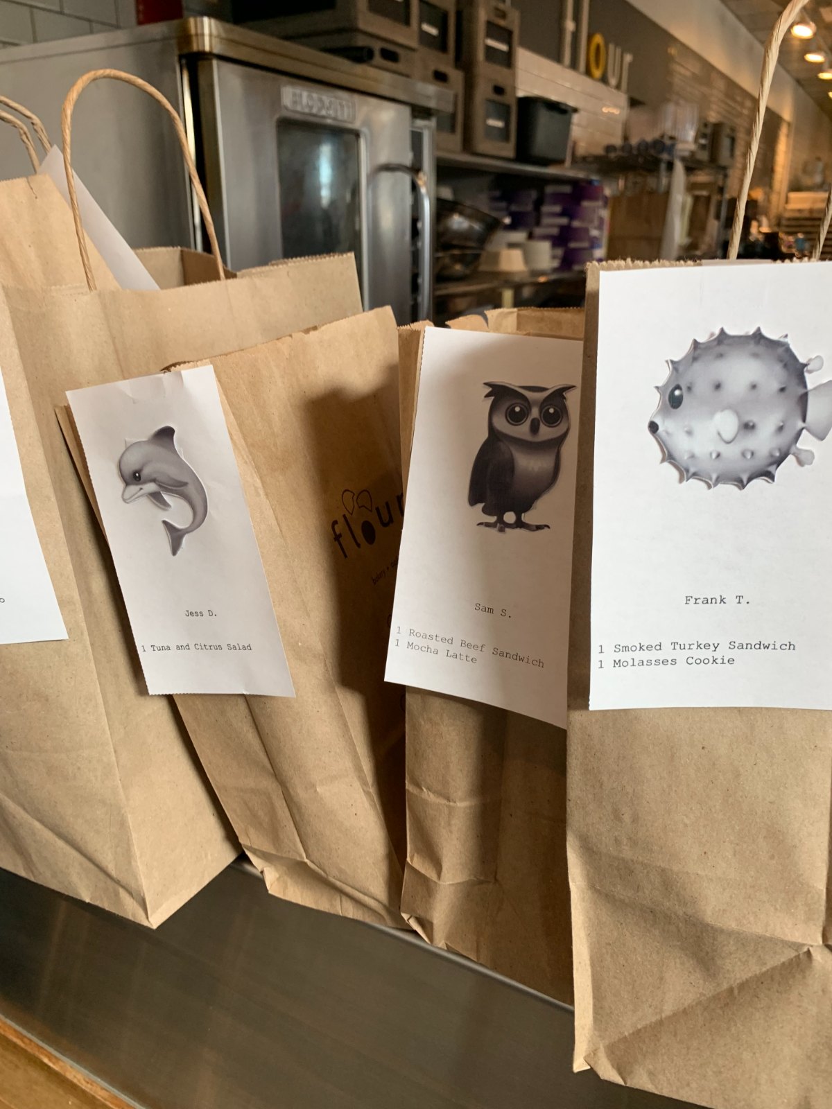 Prototype du design sprint : sacs à emporter sur un comptoir de retrait, chacun avec un ticket affichant un grand emoji (un dauphin, un hibou, un poisson-globe), le prénom et l'initiale du client, et les articles commandés